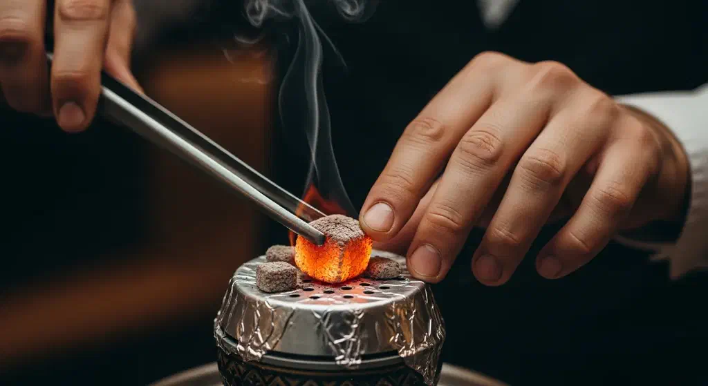 A hand using metal tongs to place a glowing coal onto the foil of a nargile pipe